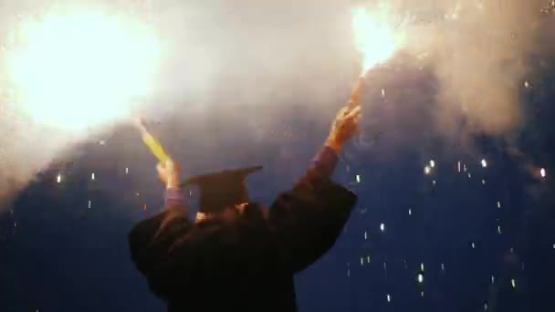 Een vrolijke afgestudeerde in een mantel en pet zwaaiend met twee vuurwerk. Afstudeerfeest — Stockvideo