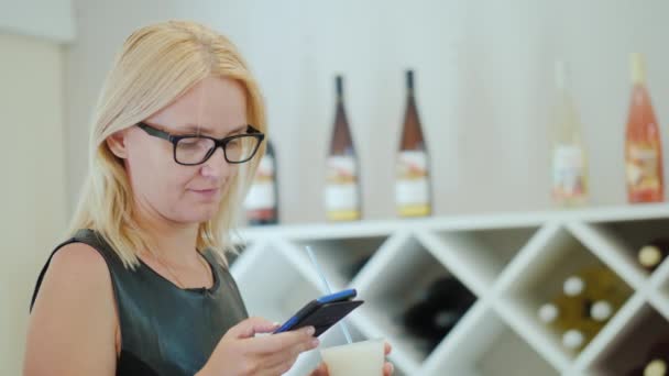 Jonge vrouw in elegante zwarte jurk maakt gebruik van een smartphone in de proeflokaal van de wijnmakerij — Stockvideo