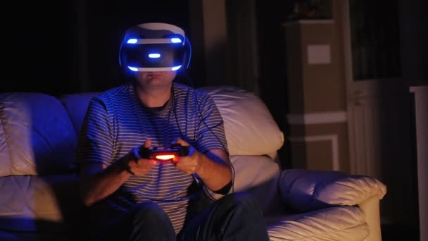 Человек играет в компьютерные игры в шлеме виртуальной реальности. Сидит на диване в темной комнате — стоковое видео