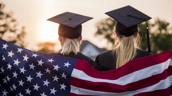 Dos graduados universitarios en batas y gorras con la bandera americana — Foto de Stock