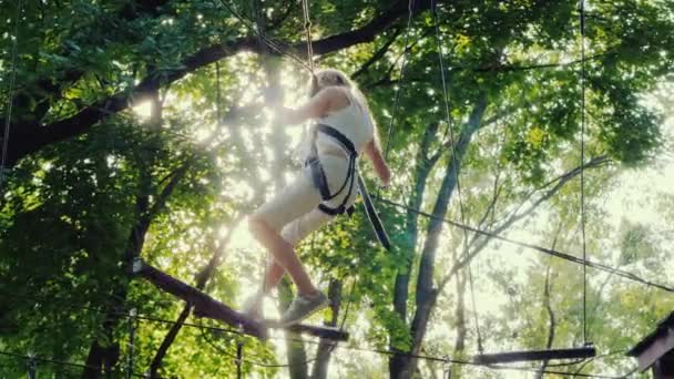 Uma criança corajosa caminha ao longo de uma corda bamba entre árvores altas. Infância ativa e diversão — Vídeo de Stock