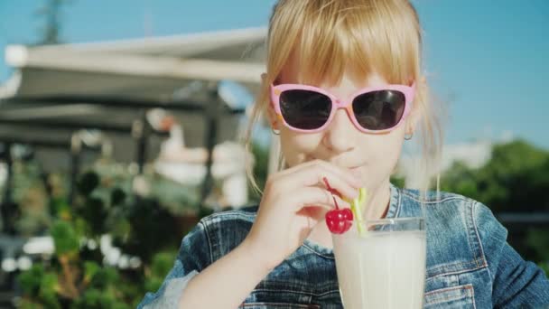 Забавная девушка в розовых очках пьет коктейль на летней террасе кафе. Отдых с детской концепцией — стоковое видео
