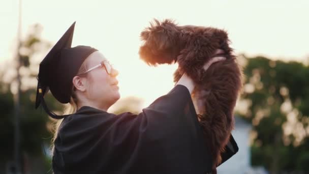 Una giovane donna in costume da laureata tiene un cucciolo carino tra le braccia. Regalo di laurea universitaria — Video Stock