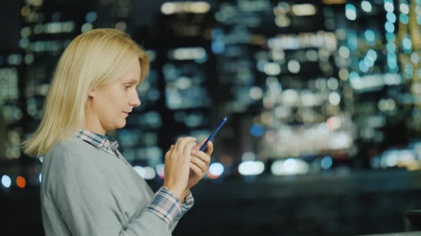 Vista lateral de la mujer blanca de mediana edad utiliza un teléfono inteligente en el fondo de las luces de Manhattan — Vídeos de Stock