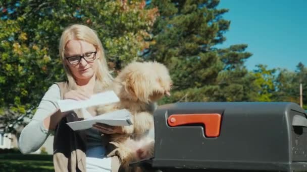 Eine junge Frau mit einem kleinen Hund auf dem Arm nimmt Briefe aus einem Briefkasten entgegen — Stockvideo