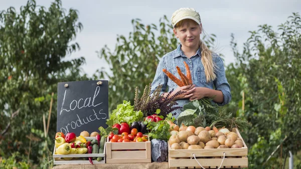 Little farmer girl at the counter with vegetables, holding a carrot — Stock Photo, Image