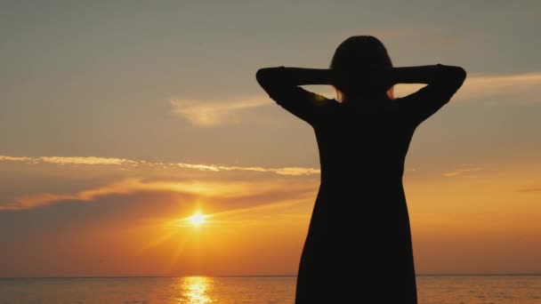 A woman admires the beautiful sky where the sun sets, stands near the sea — Stock Video