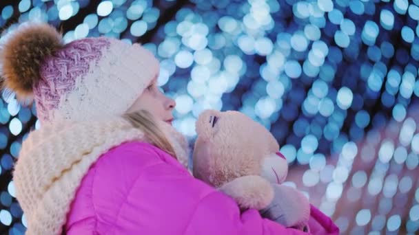 Side view of A girl with a toy in his hand looks up at the Christmas tree. — Stock Video