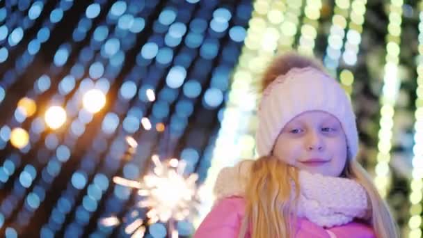 Girl playing with sparkler on the background of blurry lights of a large Christmas tree — Stock Video