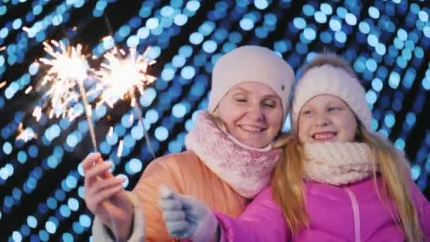 ママと娘はぼやけたクリスマスツリーライトの背景にスパークラーを再生します — ストック動画