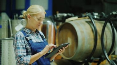 Bir kadın şaraphane atölyesinde tablet kullanır, arka planda ahşap varil vardır