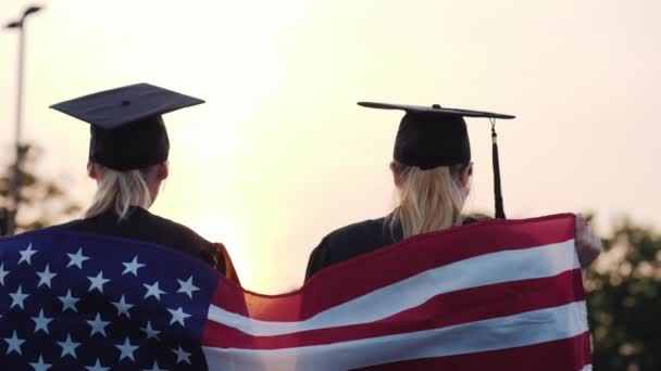 Duas mulheres graduadas universitárias levantam as mãos, sobre os ombros da bandeira dos EUA — Vídeo de Stock