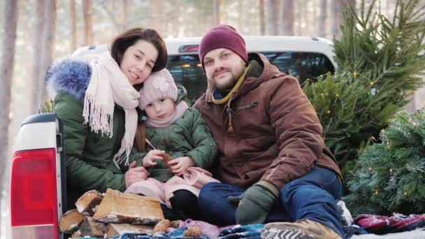 Μια ευτυχισμένη νεαρή οικογένεια ετοιμάζεται για τα Χριστούγεννα πρωί, κάθεται στο πίσω μέρος του αυτοκινήτου μετά από μια επιτυχή αγορά του δέντρου το νέο έτος, χαμογελά στη φωτογραφική μηχανή — Αρχείο Βίντεο