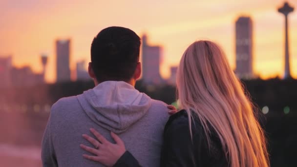 Blick von hinten auf ein junges Mädchen mit blonden Haaren, das einen asiatischen Typen auf dem Hintergrund des Sonnenuntergangs über den Niagarafällen umarmt — Stockvideo