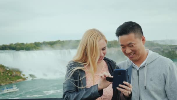 Un joven asiático y su novia caucásica están mirando una foto en un teléfono en la plataforma de observación de las Cataratas del Niágara — Vídeos de Stock