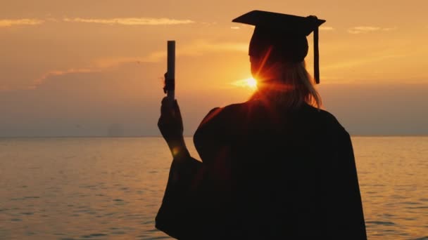 Un soltero con un diploma en la mano y una gorra de graduado mira el amanecer sobre el mar — Vídeos de Stock