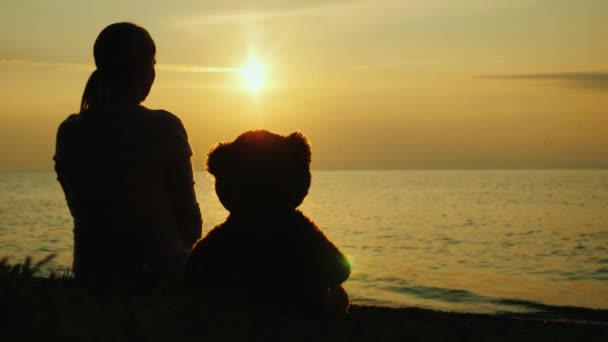 Uma mulher solitária com um urso de brinquedo olha para o pôr-do-sol sobre o mar. Conceito de tristeza e solidão — Vídeo de Stock
