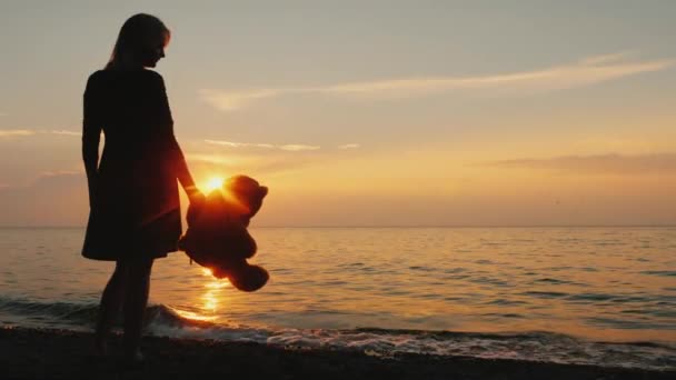 A woman with a teddy bear in hand is standing on the seashore when the sun sets. Childhood dreams — Stock Video