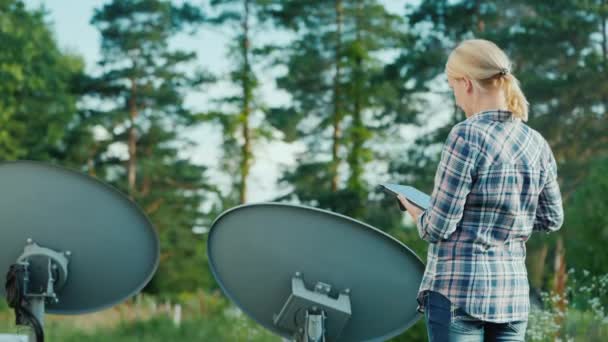 Rear view of Woman tunes satellite dishes outdoors, uses tablet — Stock Video