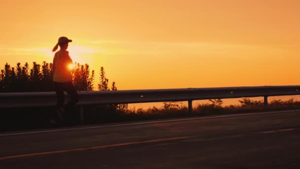 オレンジ色の空と夕日に対して道路に沿って走るスリムな運動女性 — ストック動画