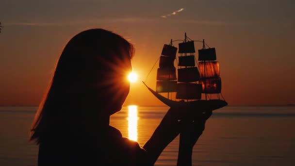 Uma mulher está à beira-mar ao pôr-do-sol, segura nas mãos um modelo de um navio à vela. Esperança e conceito de viagem — Vídeo de Stock