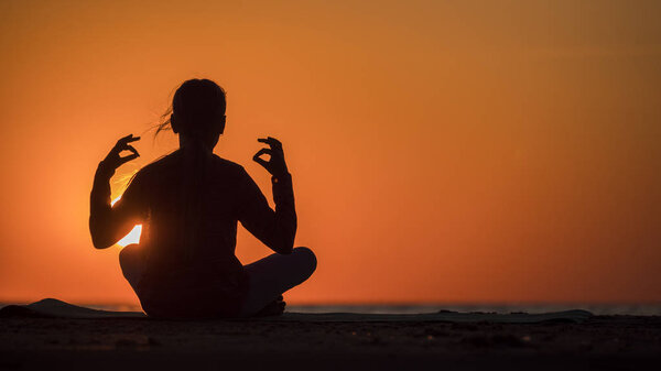 Teen girl meditating in lotus position at sunset. Sitting by the sea on a background of orange sky