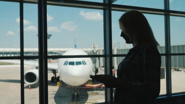 Silueta de una mujer de negocios junto a la ventana de una terminal del aeropuerto. Utiliza un teléfono inteligente — Vídeo de stock