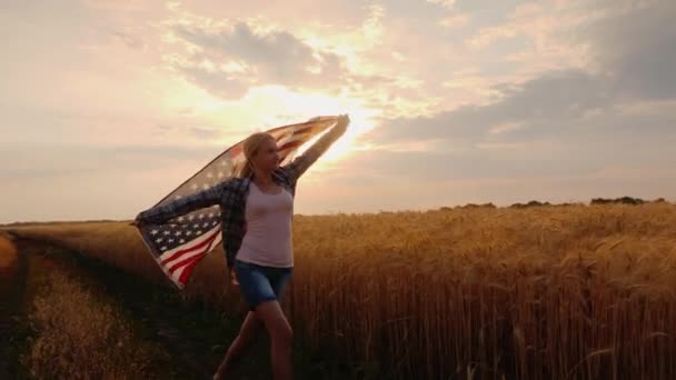 Uma mulher com uma bandeira dos EUA atravessa um campo de trigo nos raios dos sóis ao pôr do sol — Vídeo de Stock