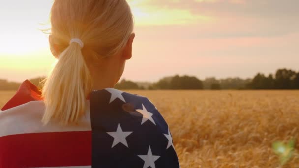 Una mujer con una bandera de EE.UU. en sus hombros disfruta de un campo de trigo al atardecer — Vídeos de Stock