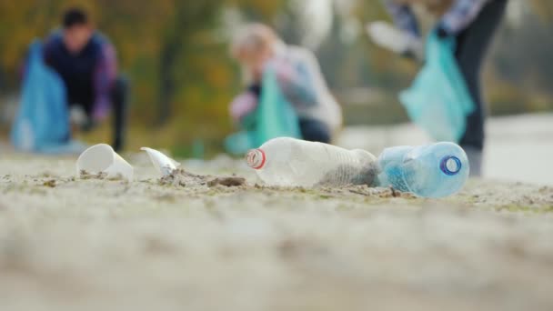 Basura de plástico en la playa, en el fondo la gente está limpiando la basura. El cuidado del concepto de medio ambiente — Vídeo de stock