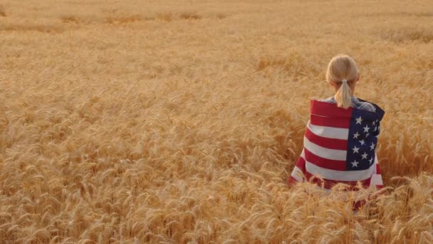 Mujer con bandera de EE.UU. sobre sus hombros está en un campo de trigo — Vídeo de stock