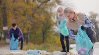 Çocuklu genç bir kadın göl boyunca çöp topluyor. Torbalara plastik atık koyun.