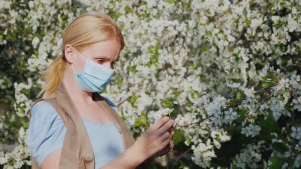 Een vrouw in een gaasverband onderzoekt een bloem tegen een bloeiende boom. Problemen met allergische aandoeningen — Stockvideo