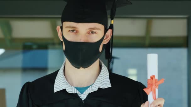 De afgestudeerde heeft een diploma in zijn hand, met een badjas, een pet en een beschermend masker op zijn gezicht. — Stockvideo