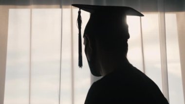 Mezuniyet şapkalı ve bornozlu mezunun dikiz görüntüsü pencereden dışarı bakıyor. Salgın sırasında koruyucu bir maske takıyor..