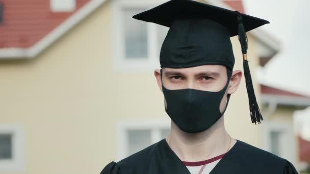 Portret van een afgestudeerde met een beschermend masker, met een badjas en een afgestudeerde pet op — Stockvideo