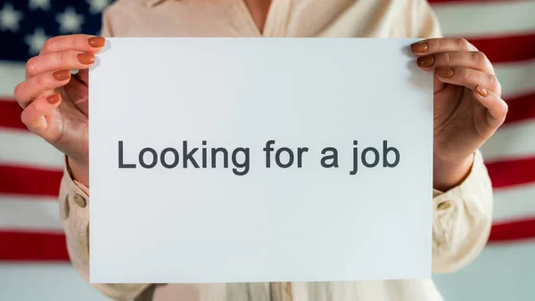 Uma mulher tem um cartaz que diz "à procura de emprego". Em pé no fundo da bandeira americana — Fotografia de Stock