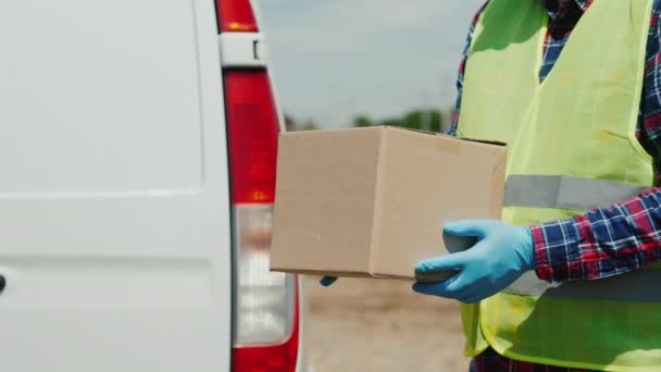 Seorang kurir dengan kotak kardus berdiri berlawanan dengan latar belakang van layanan pengiriman putih — Stok Video