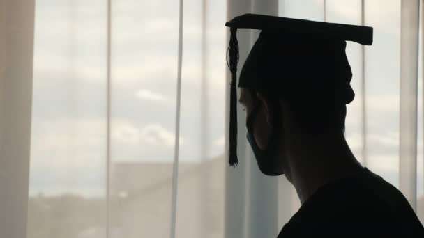 Silhouette di un laureato con cappello e mantello e una maschera sul viso. Guardando fuori dalla finestra — Video Stock