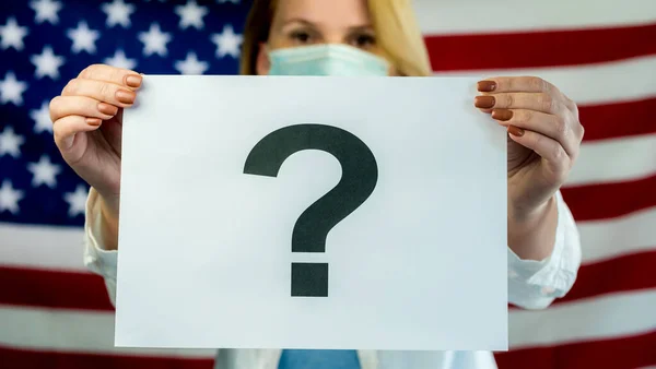 Een vrouw met een medisch masker draagt een poster met een vraagteken tegen de achtergrond van de Amerikaanse vlag. — Stockfoto