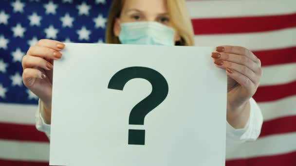 Жінка в медичній масці тримає плакат із знаком питання на тлі американського прапора. — стокове відео