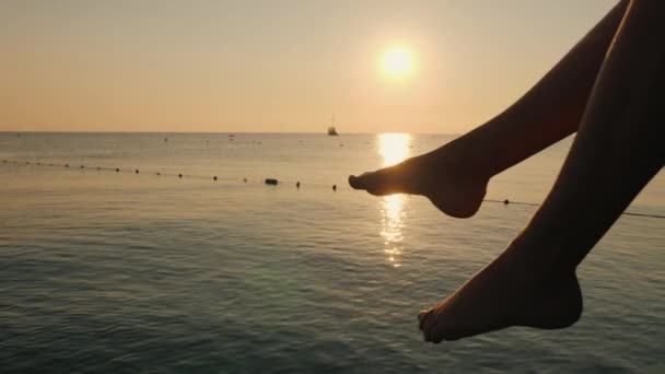 Kadın, denizin arka planında ayaklarıyla ve yükselen güneşle dikkatsizce sohbet ediyor. Uzakta bir gemi görülüyor. Sadece çerçevedeki bacaklar. — Stok video