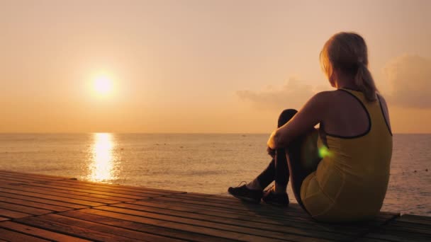 Een jonge vrouw zit op een pier en bewondert de zonsopgang boven de zee. — Stockvideo