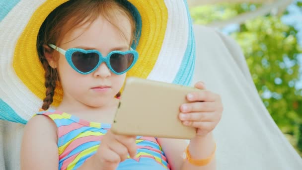 En la playa con un smartphone. Niña está jugando en el teléfono inteligente, está sentado en un gran sombrero en chaise — Vídeo de stock