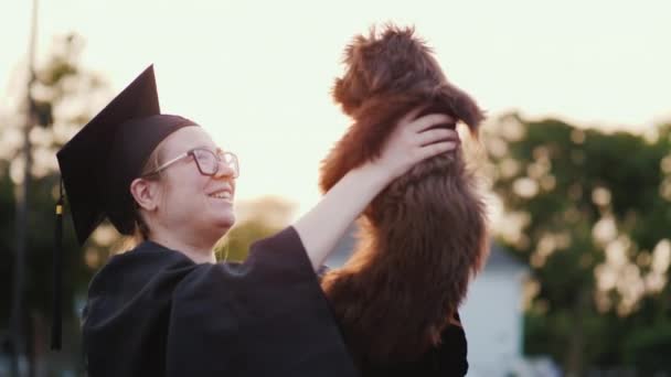 Regalo para la graduación: una estudiante con un manto y una gorra sostiene a un cachorro en sus manos — Vídeo de stock