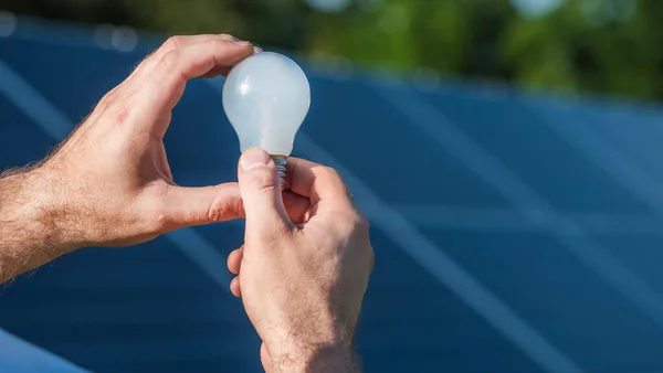 Las manos de Mans sostienen una bombilla contra el fondo de los paneles solares — Foto de Stock