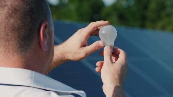 Un homme regarde une ampoule électrique sur fond de panneaux de centrales solaires — Video
