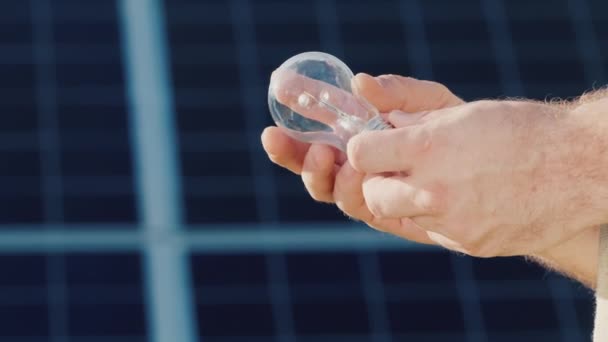 Вид збоку: руки в руках тримають лампочку на фоні сонячних батарей. — стокове відео