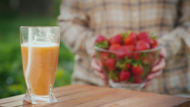 農家はジュースのガラスの横に、新鮮なイチゴと透明なボウルをテーブルの上に置きます — ストック動画