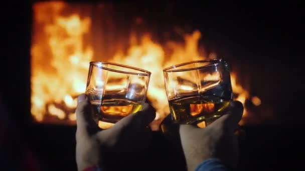Due mani maschili con bicchieri di whisky con ghiaccio sullo sfondo del camino. Concetto di fuga invernale — Video Stock